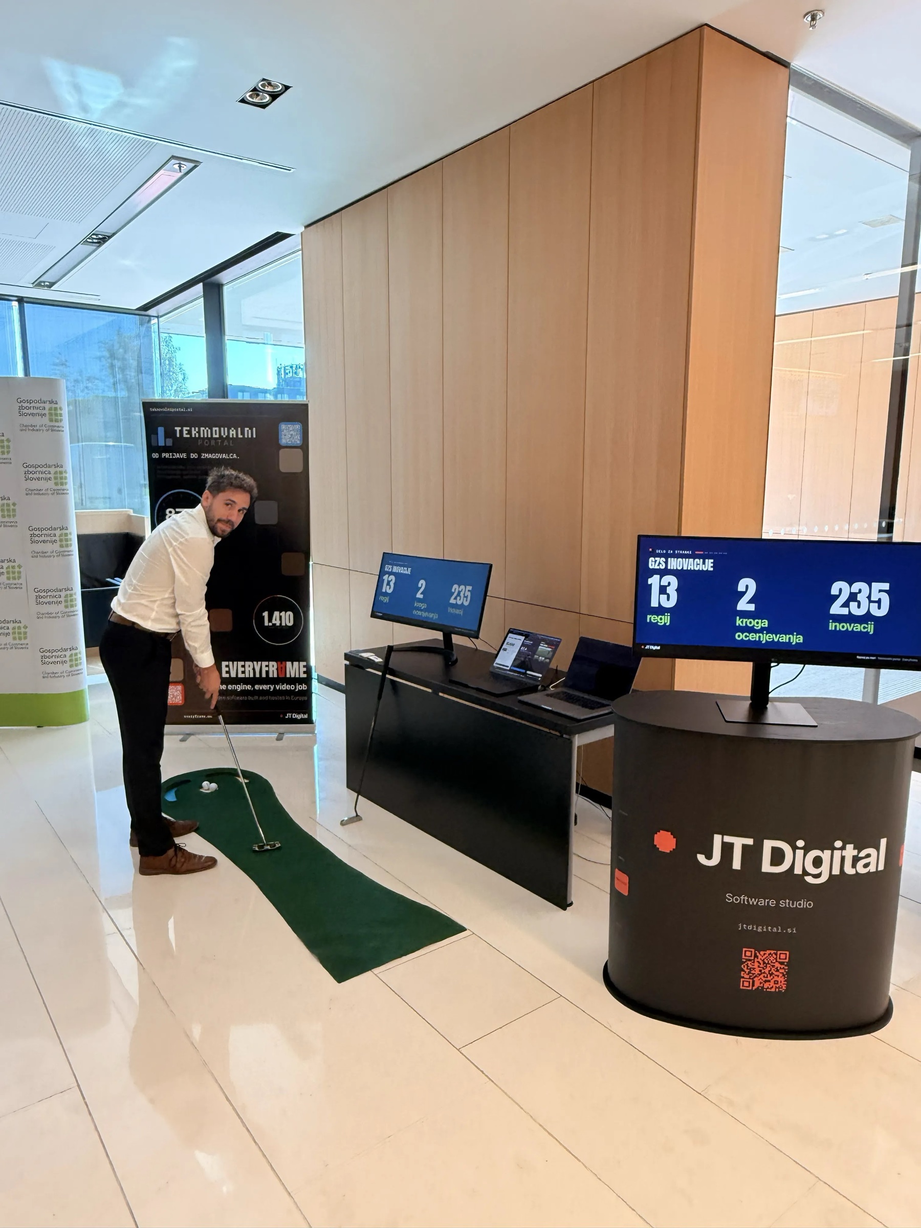 A JT Digital team member putting on the mini golf green at the JT Digital stand, with GZS Inovacije results on screen: 13 regions, 2 rounds, 235 innovations